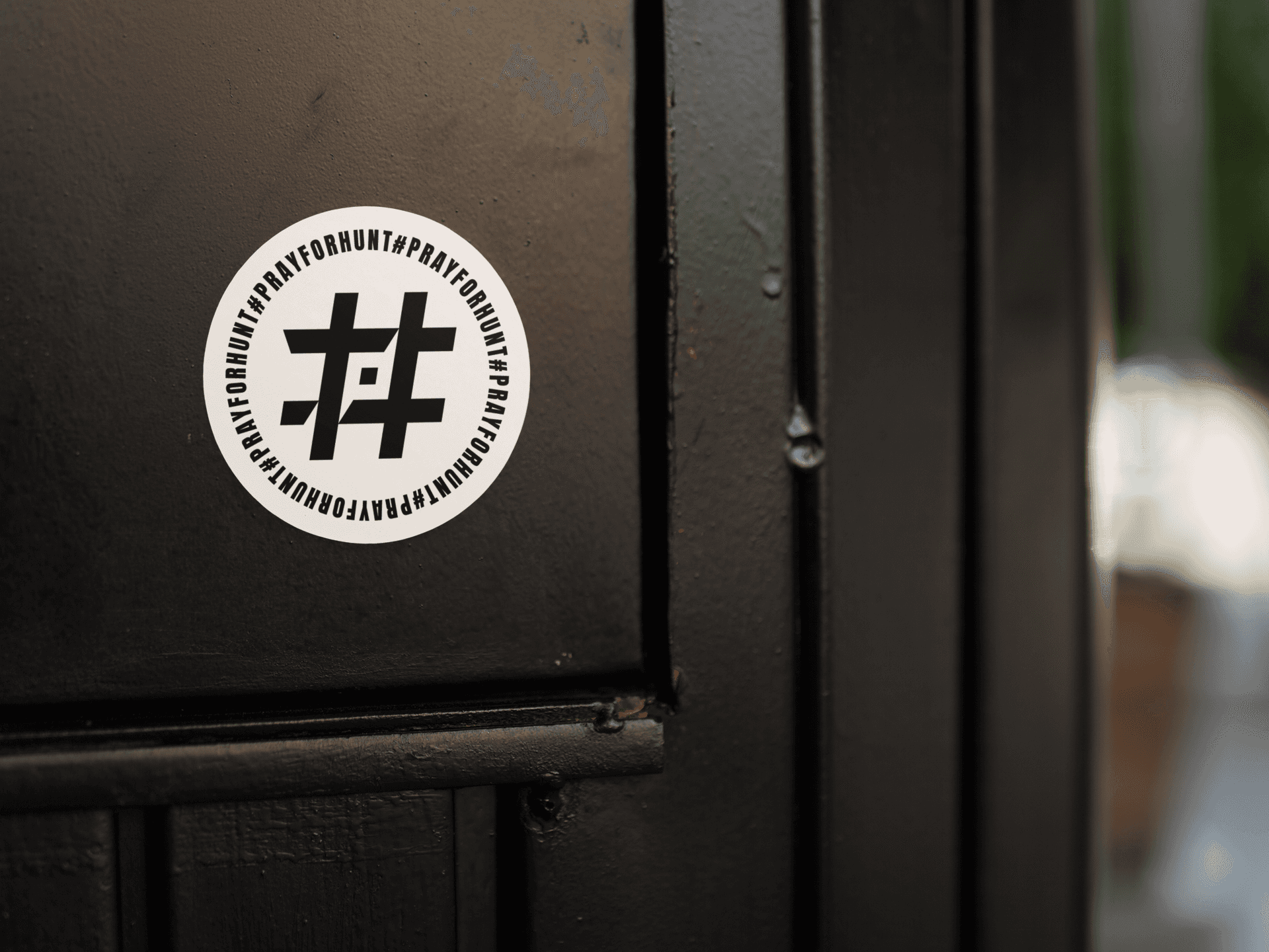 A round sticker featuring the '#PrayForHunt' logo applied to a black, textured surface, likely a door. The sticker is white with a bold, black hashtag symbol in the center that incorporates a Shuriken element. Surrounding the symbol, the phrase '#PrayForHunt' repeats in a circular pattern, emphasizing the message. The background shows subtle reflections and depth, highlighting the sticker's placement and making it stand out prominently.