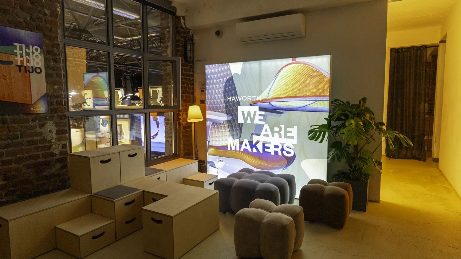 We Are Makers light wall with grey poufs and plywood box seating at the Haworth Orgatec booth.
