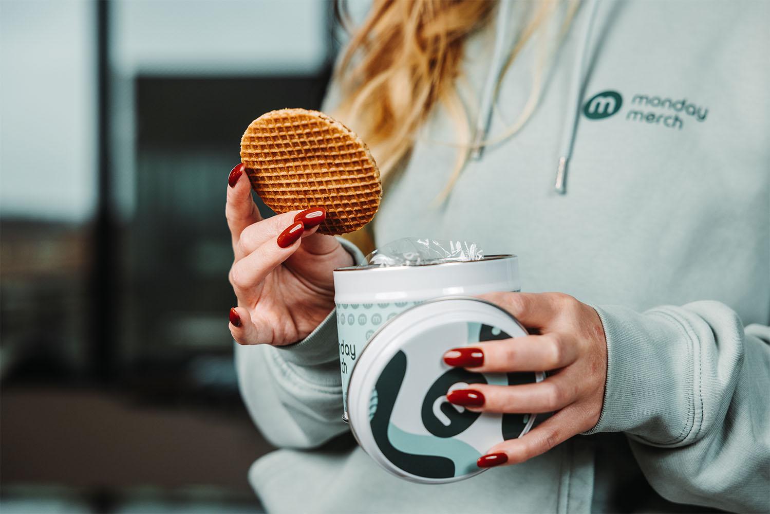 Personne tenant un stroopwafel et une boîte ouverte, portant un hoodie avec le logo "monday merch"