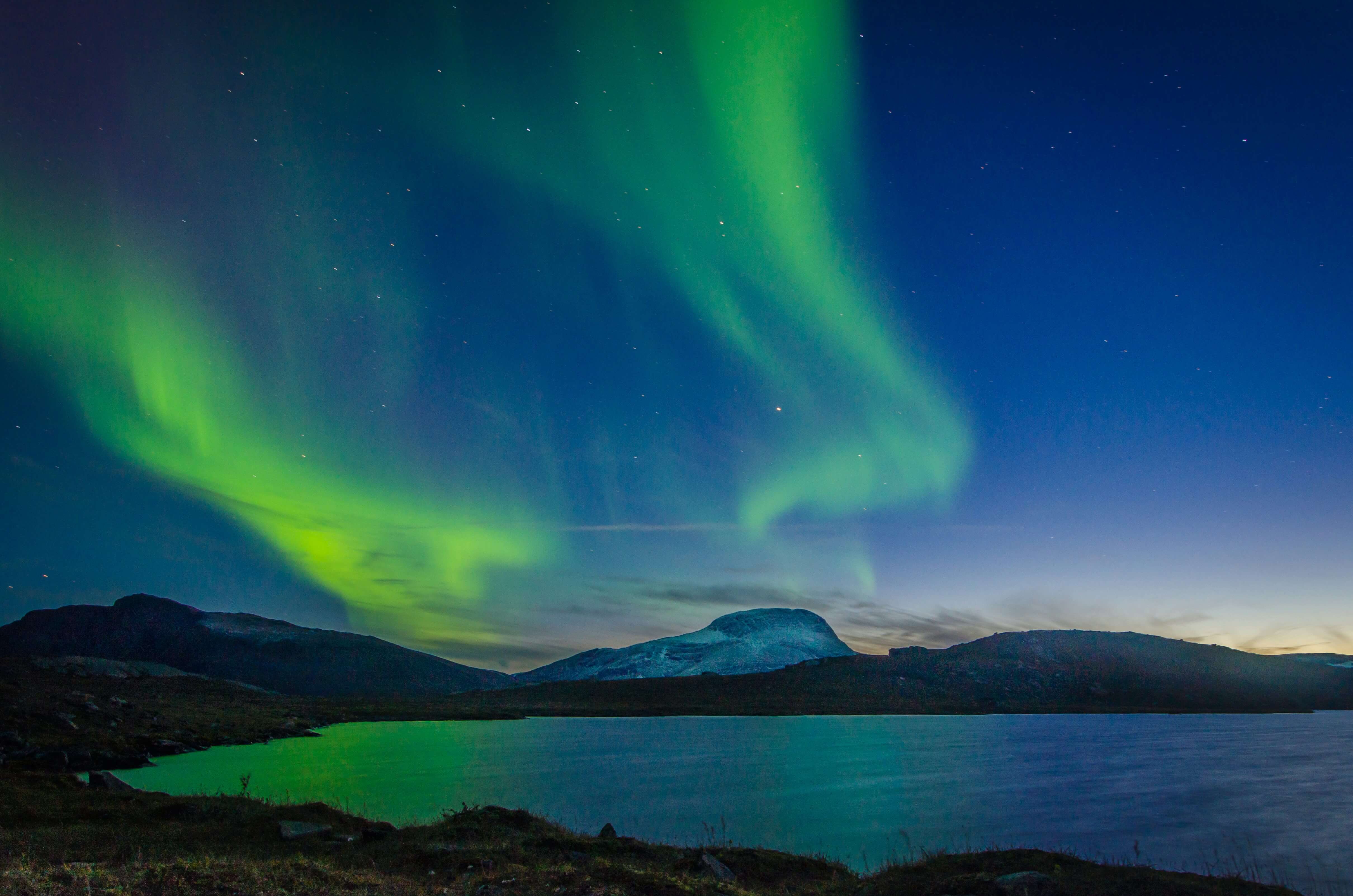 This stunning image captures the Northern Lights (Aurora Borealis) dancing across the night sky over a serene landscape. The vivid green auroras swirl and stretch across the deep blue and purple hues of the sky, reflecting beautifully on the calm water below. Snow-capped mountains rise in the distance, adding depth and grandeur to the scene. The photo conveys a sense of awe and tranquility, highlighting the natural beauty and wonder of the Arctic or sub-Arctic region where this phenomenon occurs—likely in parts of Norway, Sweden, Finland, Canada, or Alaska.