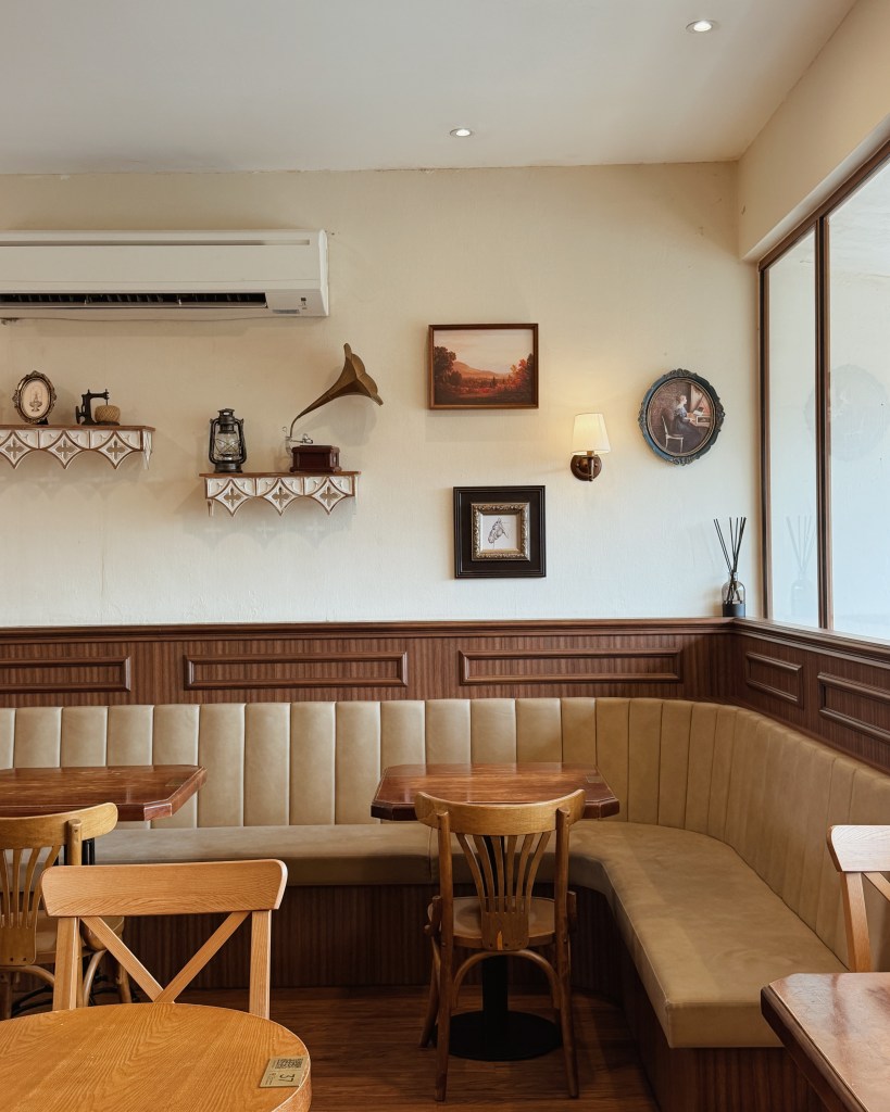 A cozy café interior featuring wooden tables and chairs, a beige upholstered bench, and vintage decor items on the wall, including paintings and a gramophone.