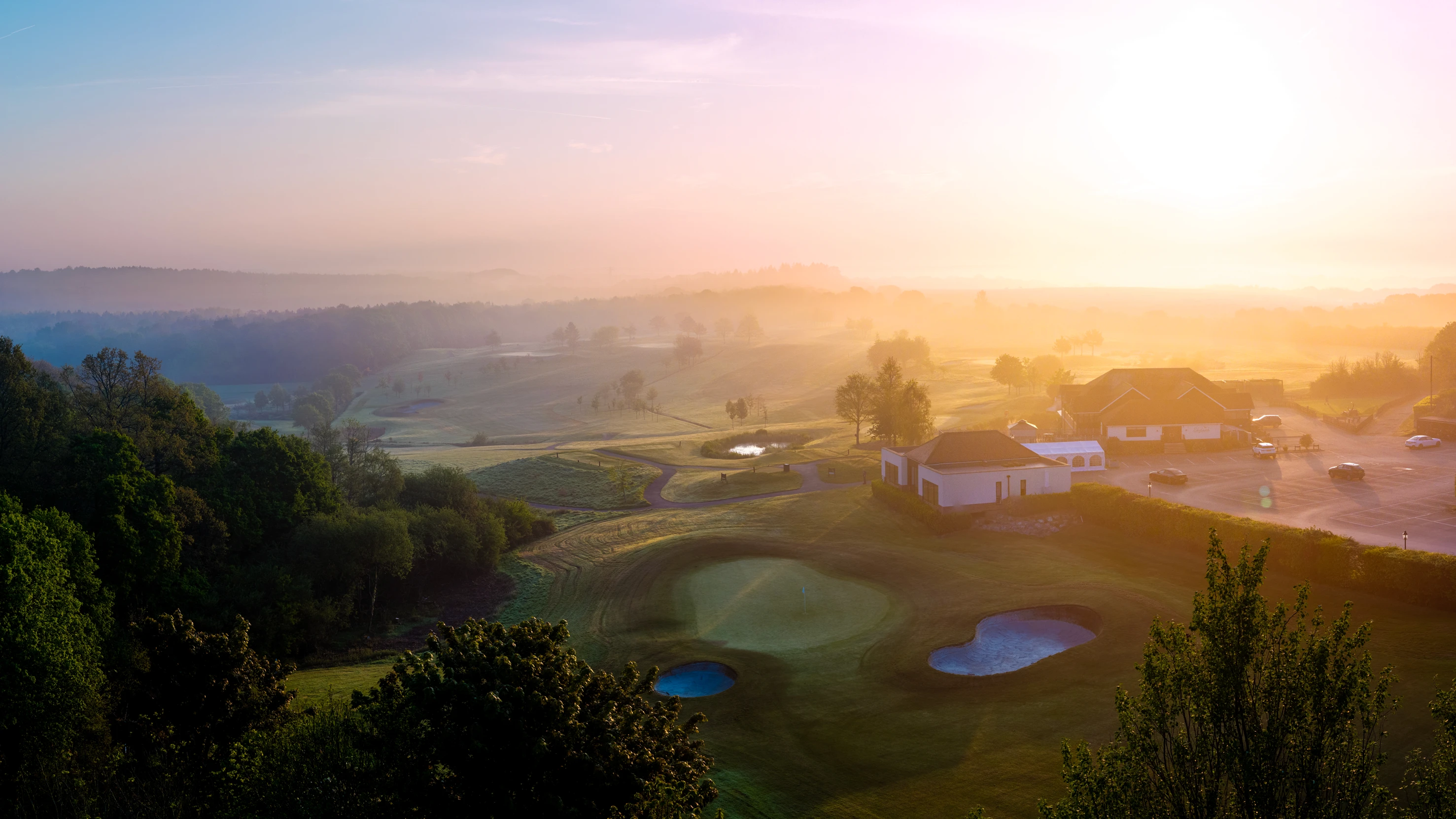 Golf course at sunrise with a clubhouse and parking lot.