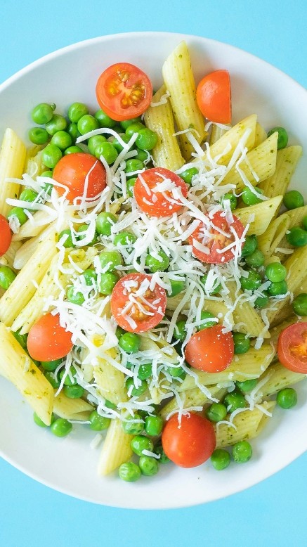 Plate of vegetarian pesto penne with fresh tomatoes garnished with peas, and cheese on a blue background