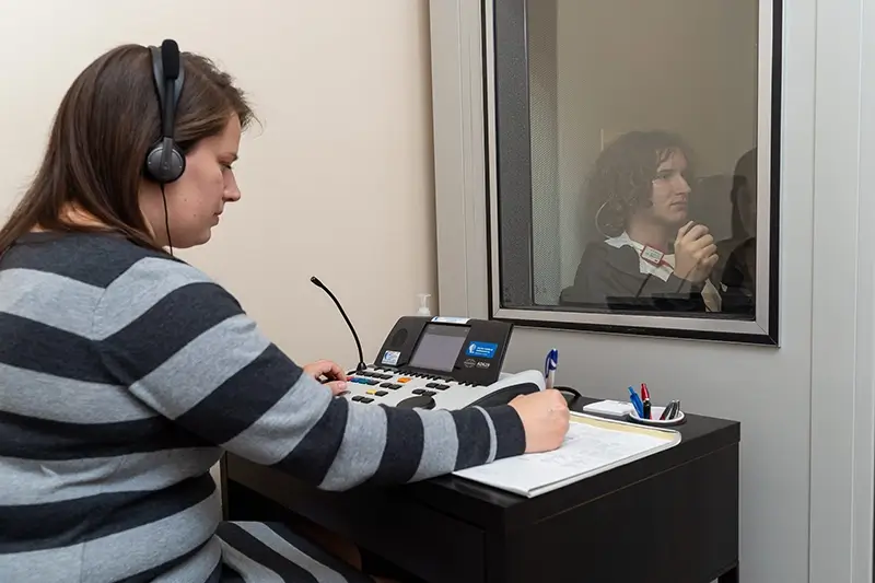 A person wearing headphones sits at a desk, observing someone behind a glass partition.