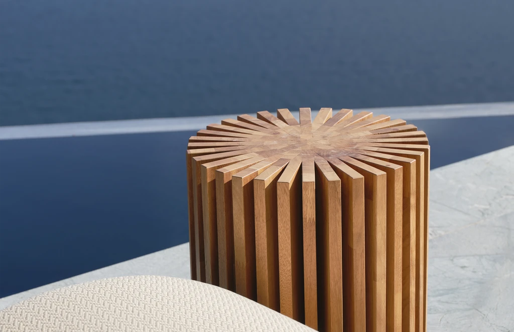 Round outdoor side table in solid Iroko wood, crafted with interlocking vertical panels in a radial pattern to form a sculptural cylindrical shape – featured poolside in a luxury modern setting with lounge chairs.