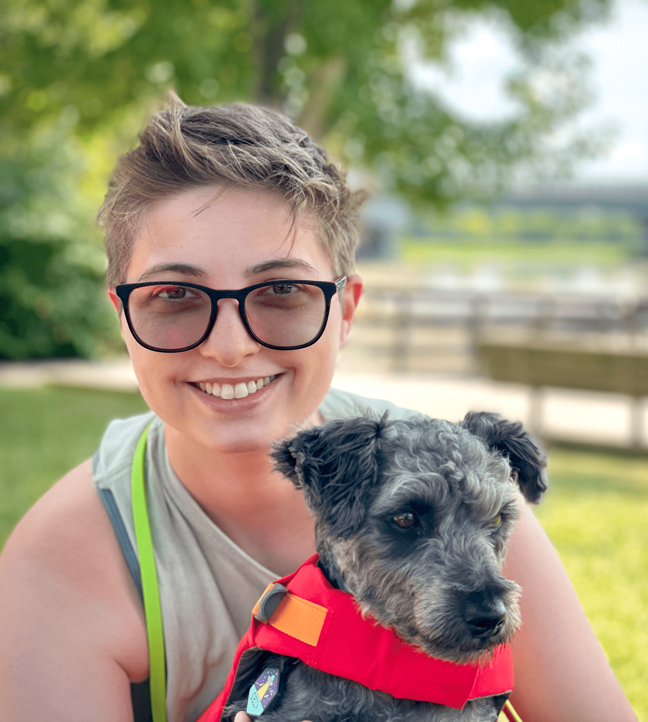 An image of Yasmine and her dog, a black & gray schnoodle named Gibson. Yasmine has short hair, is wearing transition lenses, and a tank top. Gibson is wearing a doggie life vest. They are about to go kayaking, and the background is the kayaking area, a park with a lake. 