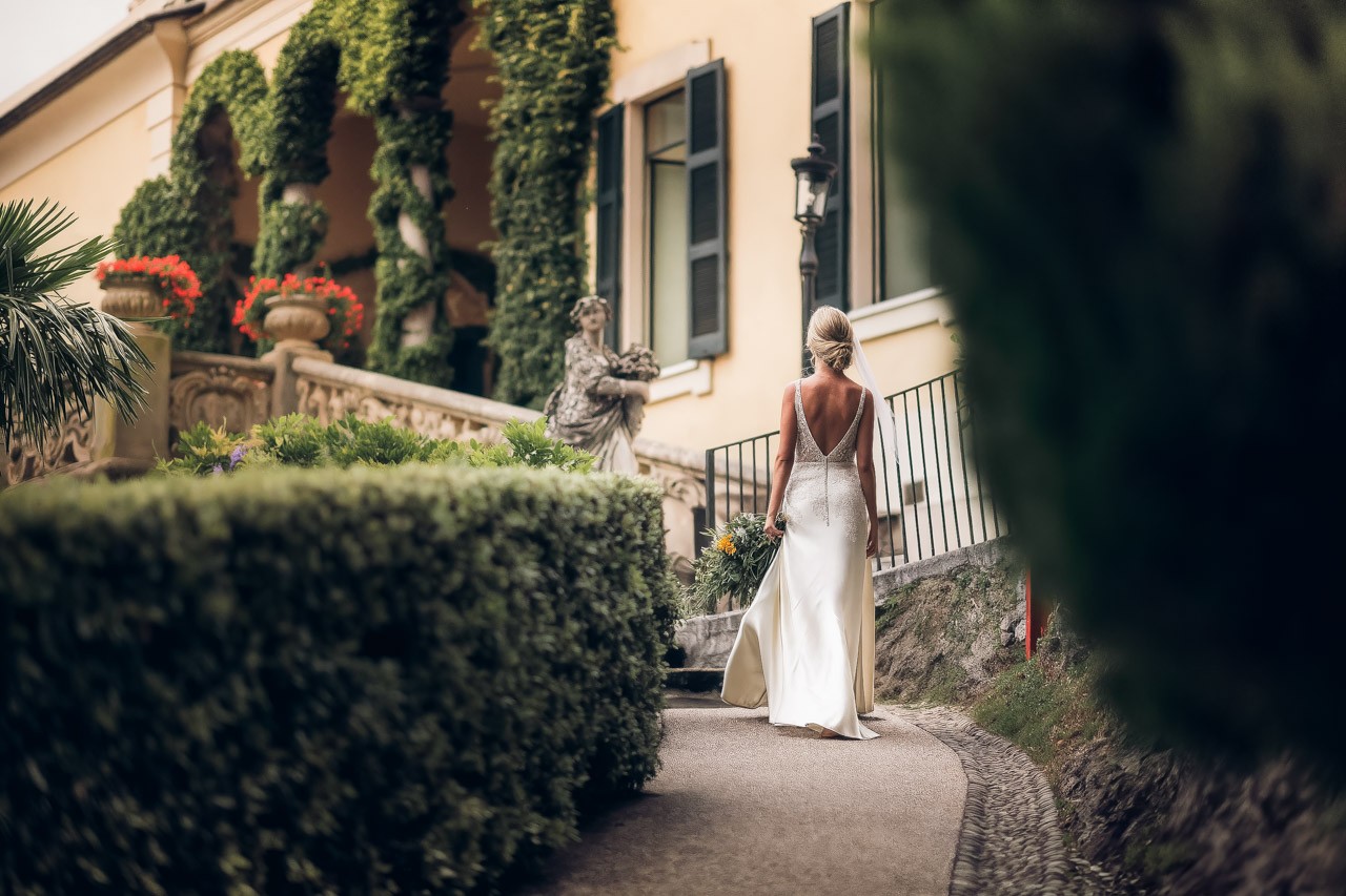 A woman in a white dress walks along a pathway surrounded by lush greenery and a charming stone building.