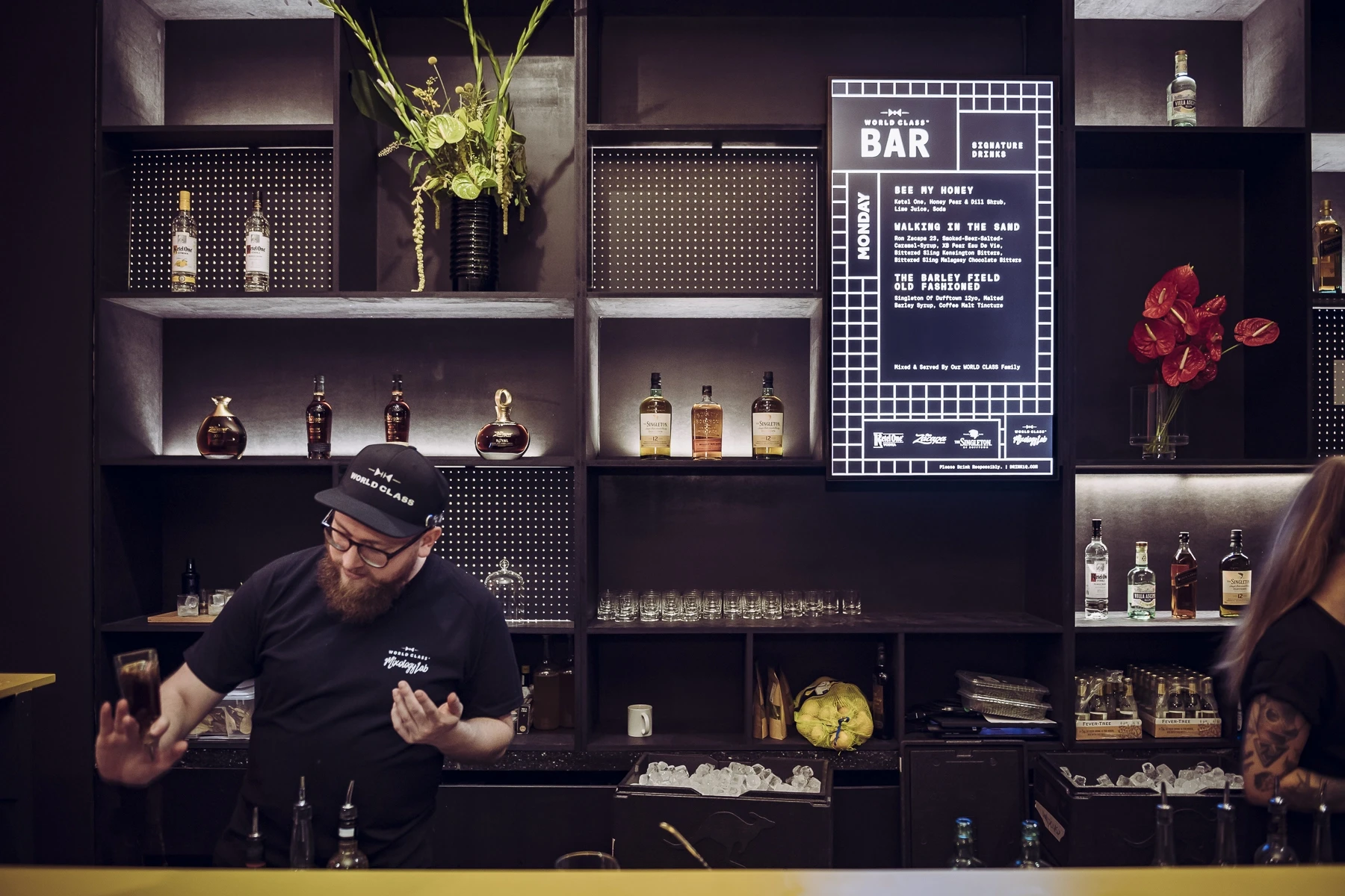 Bartender behind the World Class bar at DIAGEO BCB19 with black shelving, spotlit bottles, flowers and an illuminated grid menu board.