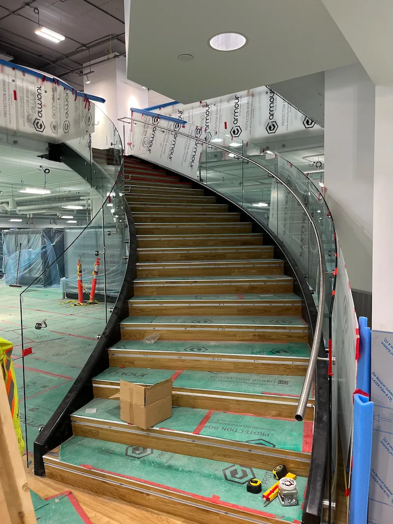 Curved glass staircase under installation inside a commercial building, showing custom metal handrail and precise structural detailing.