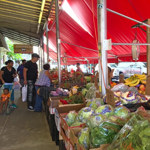 Outdoor market with red canopies displaying assorted vegetables and fruits. People are browsing and shopping.