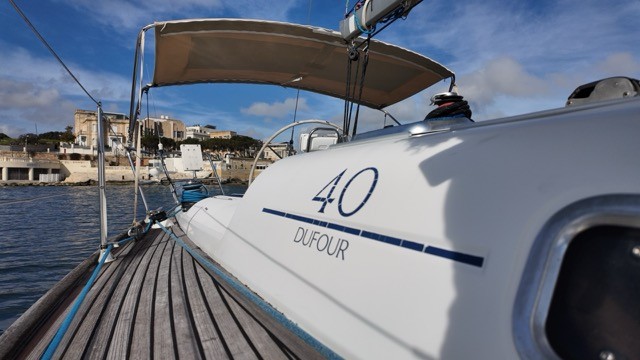 Close-up view of the Dufour 40 Performance model name on the white hull, with the number 40 and Dufour lettering in navy blue, teak side deck visible in the foreground, and a Maltese limestone waterfront with historic buildings under a blue sky in the background
