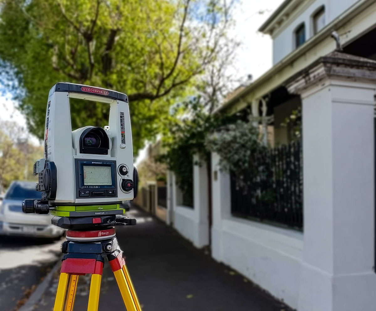 Surveying total station set up on a tripod beside a residential street with houses in the background.