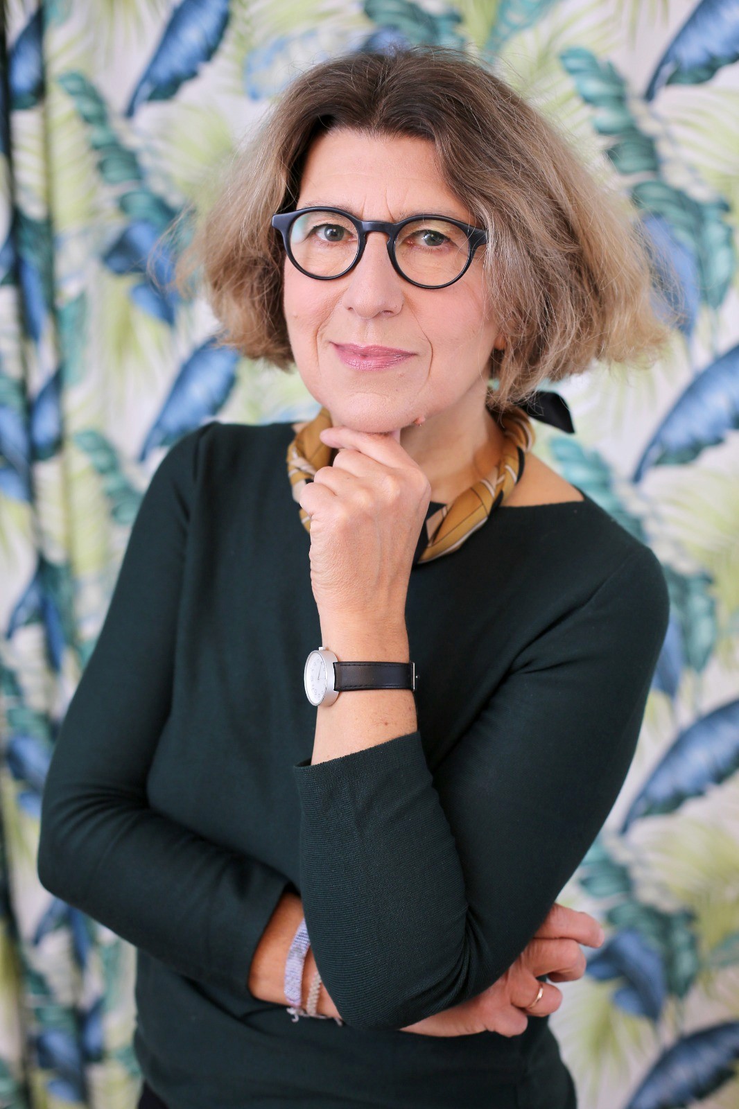 Smiling female therapist with short gray hair, wearing a brown top and a purple shawl, surrounded by a cozy background.