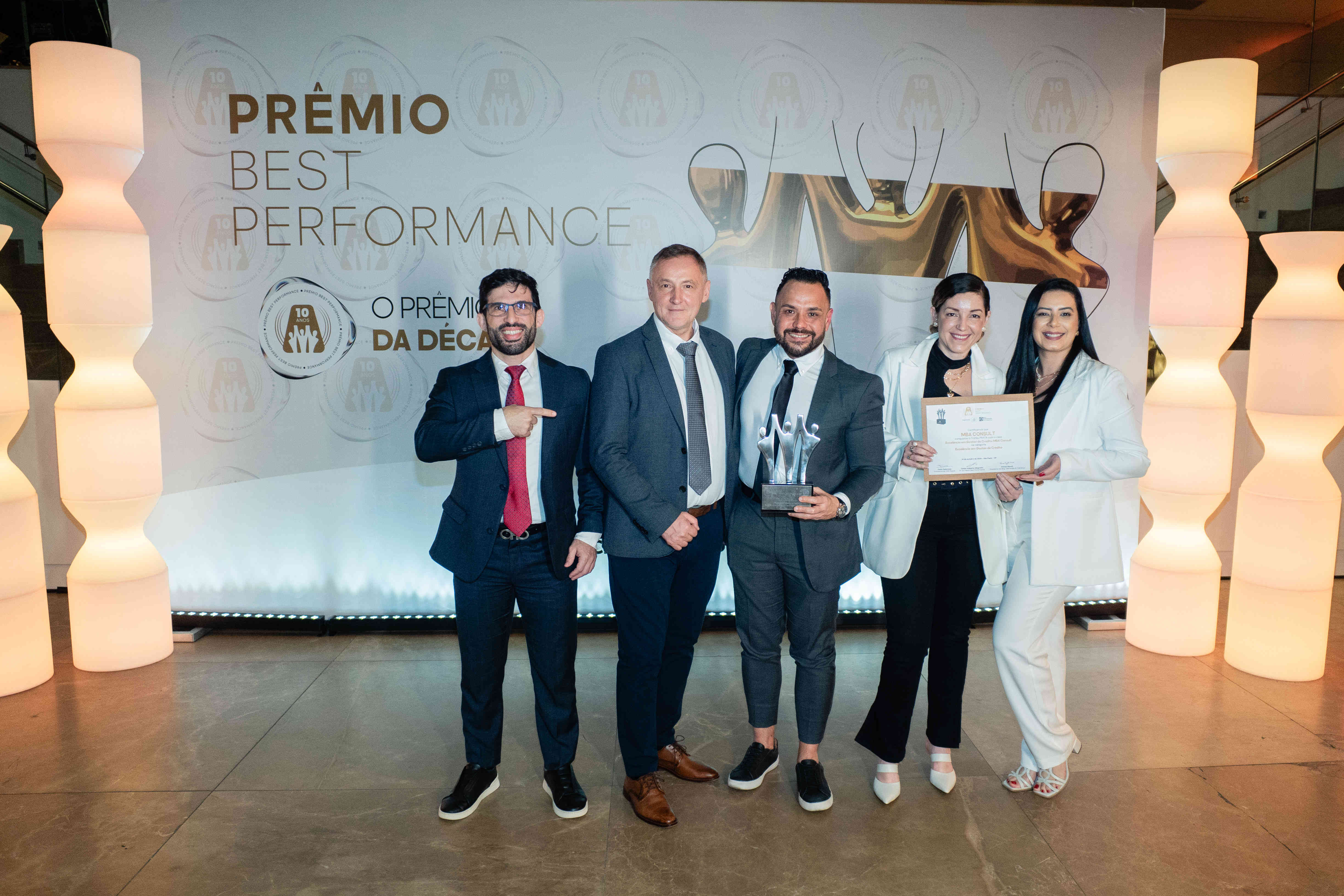 Executivos da MBA CONSULT posando com troféu prata e certificado no palco do Prêmio Best Performance 2025