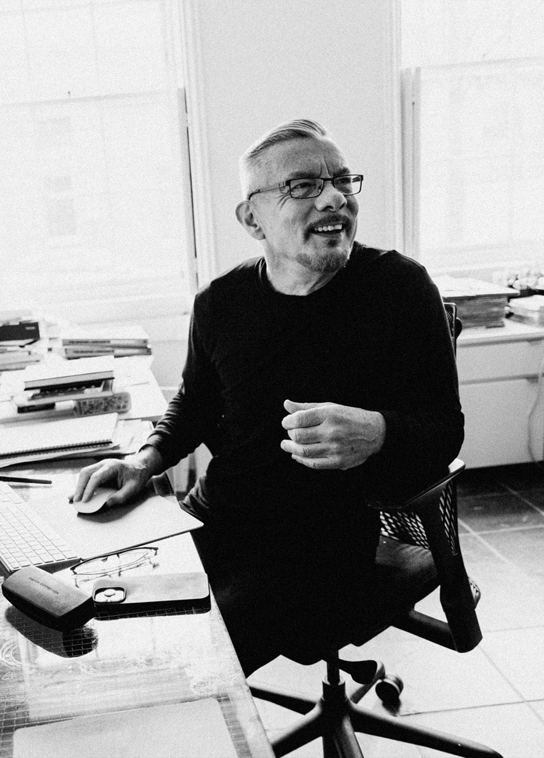 Neville Brody in a black outfit sits in an office, gesturing while engaged in conversation or thought.