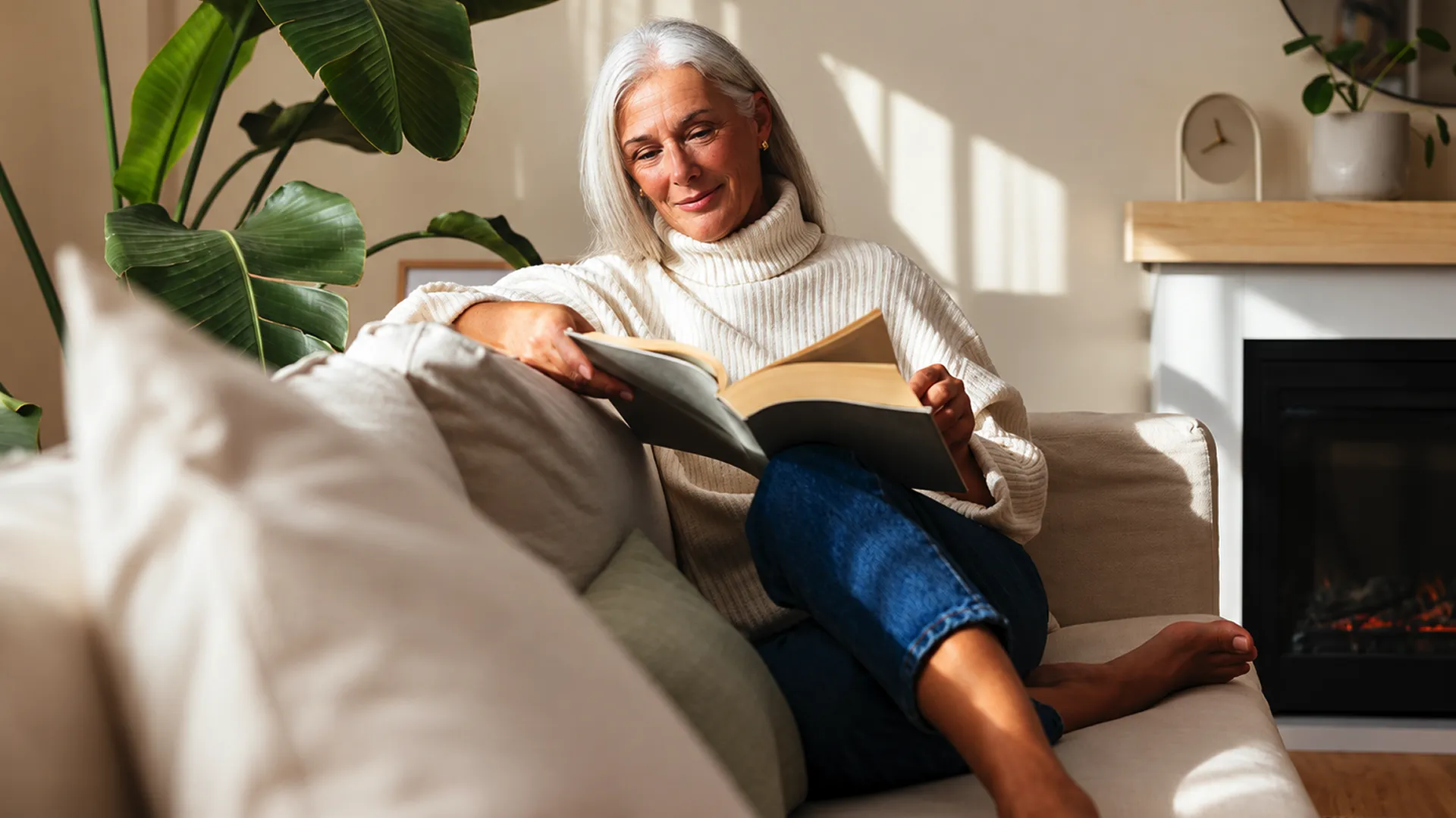 Middle-aged woman sitting on a couch and reading a book, representing vitality, longevity, and the power of preventive care.