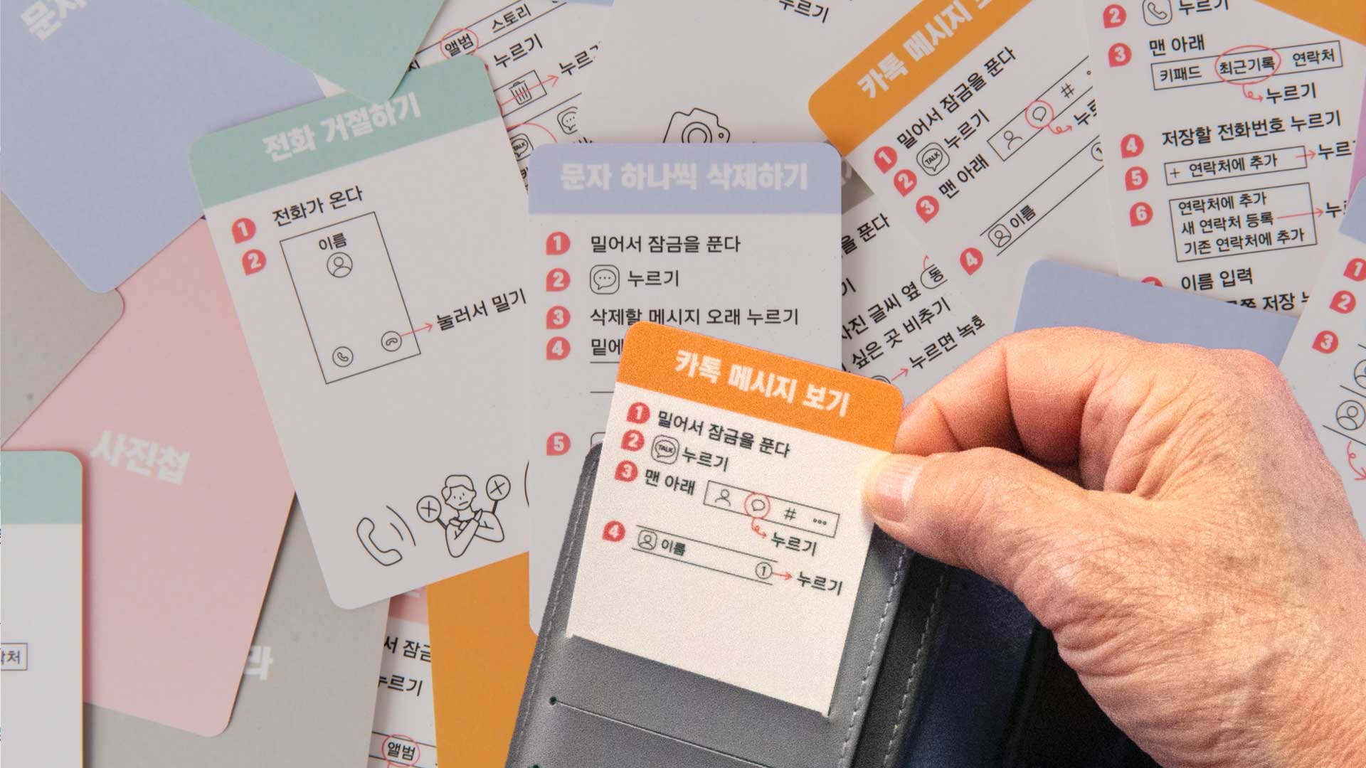 A hand holds a card with an orange top section displaying Korean text and a white bottom section with black text. The card is partially inserted into a black wallet. Surrounding the card are multiple other cards with similar designs, some featuring Korean text and simple drawings. The cards are scattered and overlap each other.