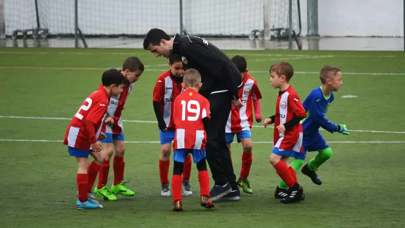 A youth football coach in a black jacket is seen engaging with a group of enthusiastic young players wearing red and blue jerseys during a funino tournament. This image captures the essence of the child football reform, where the focus is on active participation, skill development, and enjoyment, which are core elements of kids' festivals.