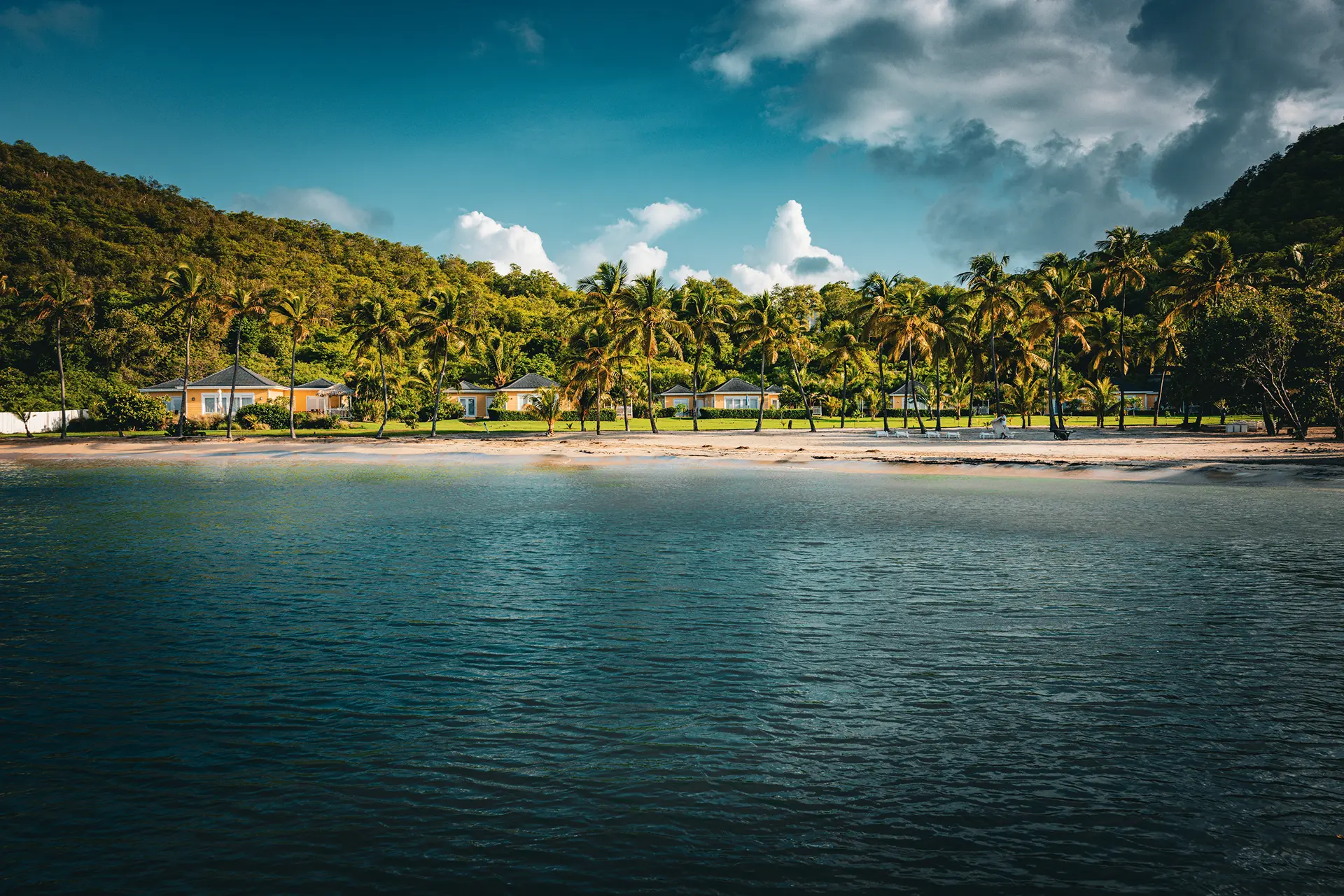 Golden-hour view of a tranquil beachfront retreat with calm ocean water, thatched-roof pavilion, and lush hillside backdrop