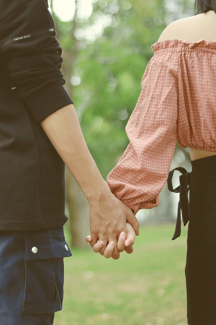 Close-up of two people holding hands. The person on the left wears a black long-sleeved shirt with "CHICAGO BULLS" printed on the sleeve and navy blue cargo pants. The person on the right wears an off-the-shoulder, coral-colored checkered shirt with puffed sleeves and a black skirt with a tie. They stand outdoors with a blurred green background.