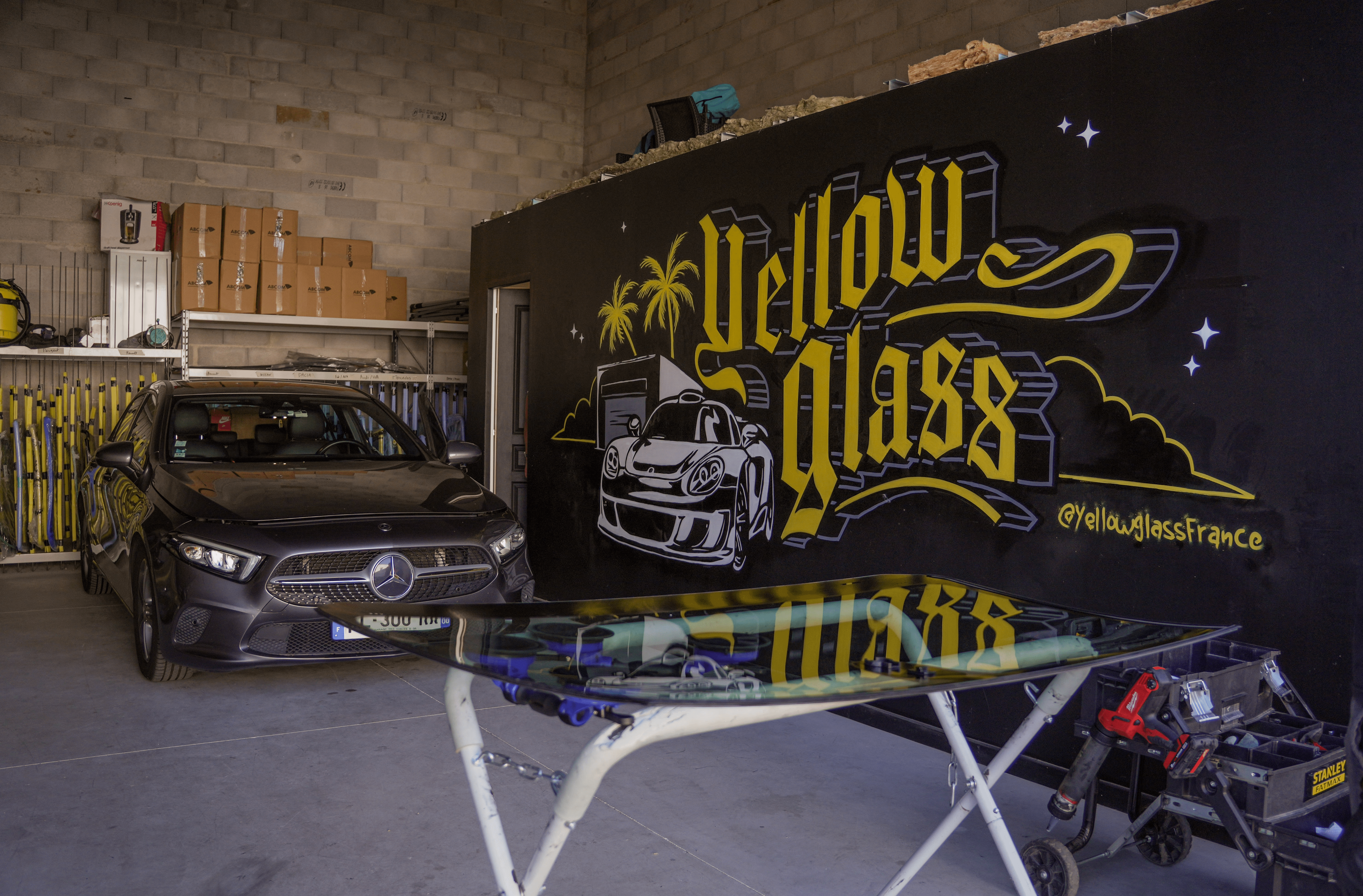 Une agence de remplacement de pare-brise avec une voiture Mercedes noire devant un mur avec un logo "Yellow Glass". Réparation automobile professionnelle.