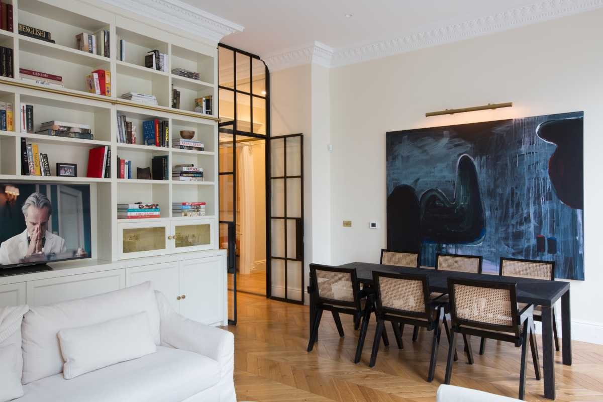Open-plan dining and living space designed by Doll & Co. Islington Architects, featuring custom floor-to-ceiling built-in shelving, black Crittall-style doors, and abstract art