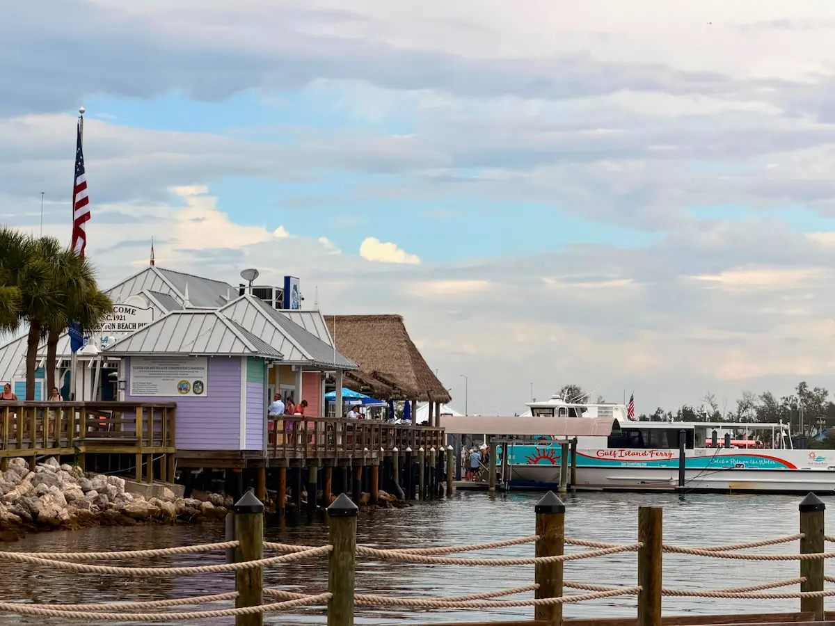 A scenic view of a quaint waterfront marina features colorful buildings with a thatched roof, a docked tour boat labeled "gulf islands ferry," docked at historic Bridge Street pier on Anna Maria Island
