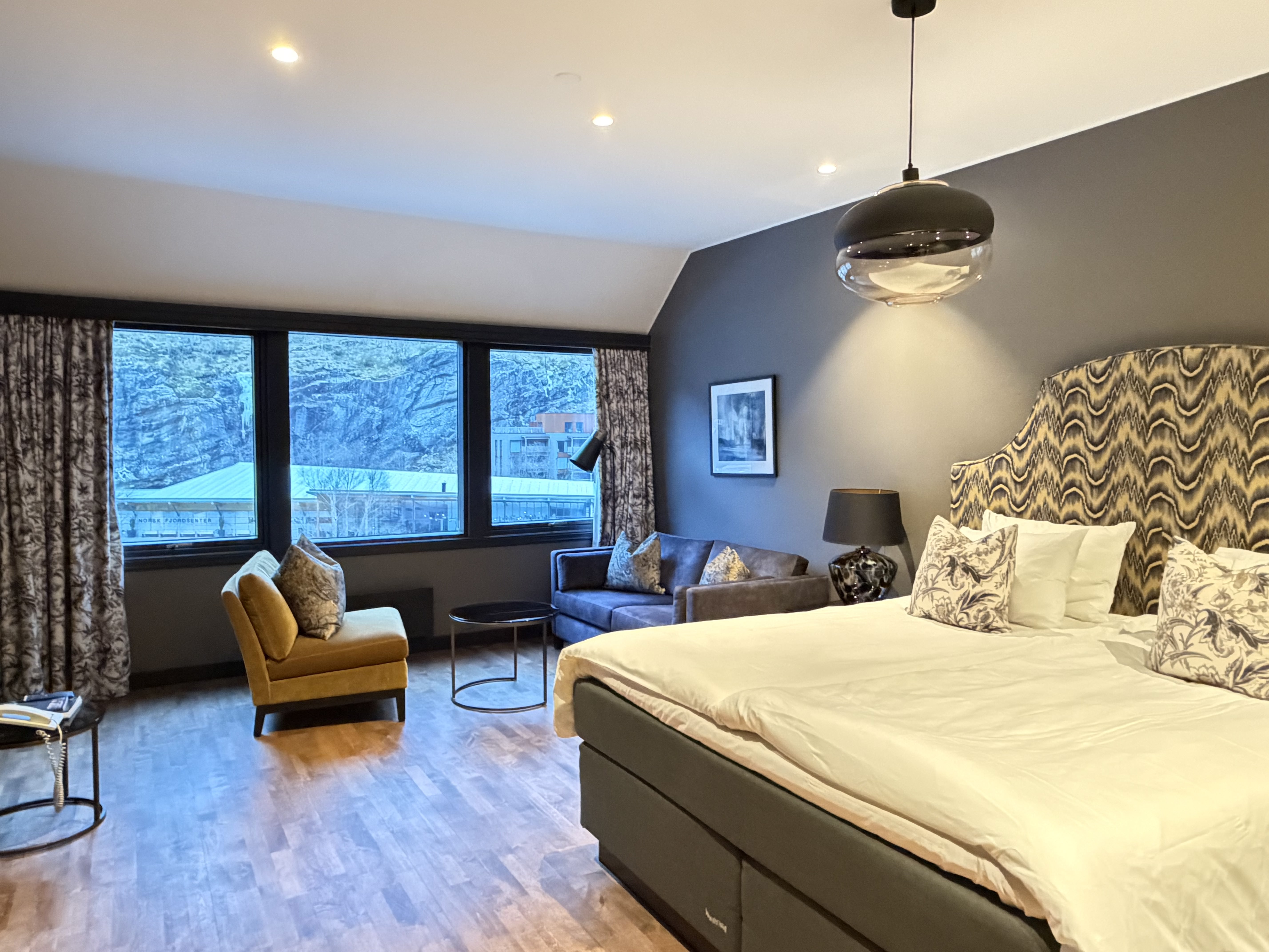 A cozy hotel room features a large bed with green and white bedding, set against a wooden accent wall, with a modern pendant light above, a blue sofa near a large window offering a scenic outdoor view, and a work desk with a mounted TV.