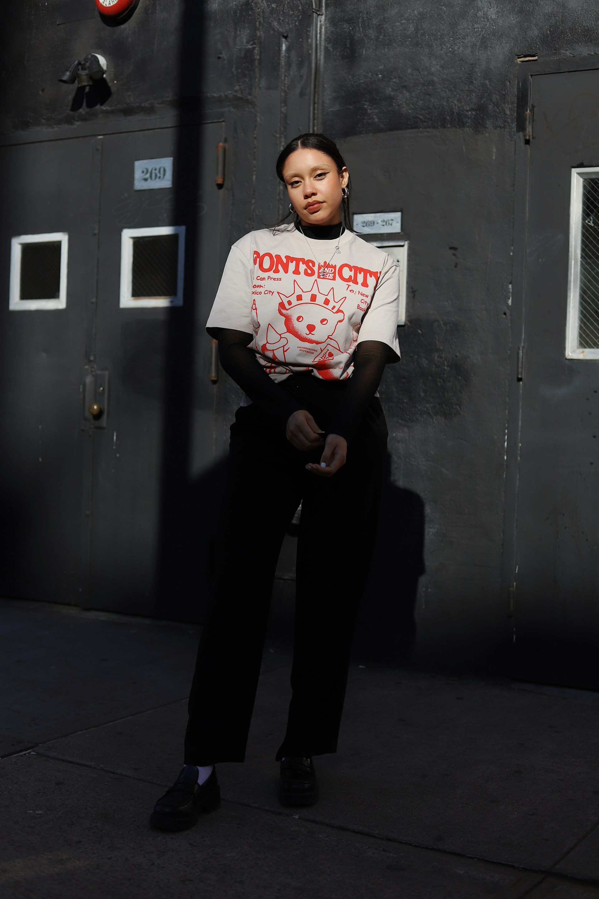A full-body shot of the woman standing against a dark grey industrial building with heavy metal doors. She is wearing a grey graphic t-shirt, black mesh sleeves, black trousers, and black loafers.