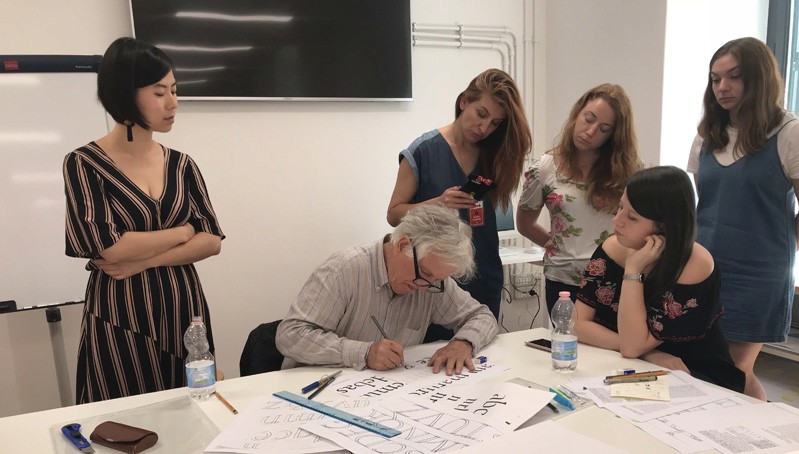 A photo showing students looking at a teacher that is explaining how to draw text letters.