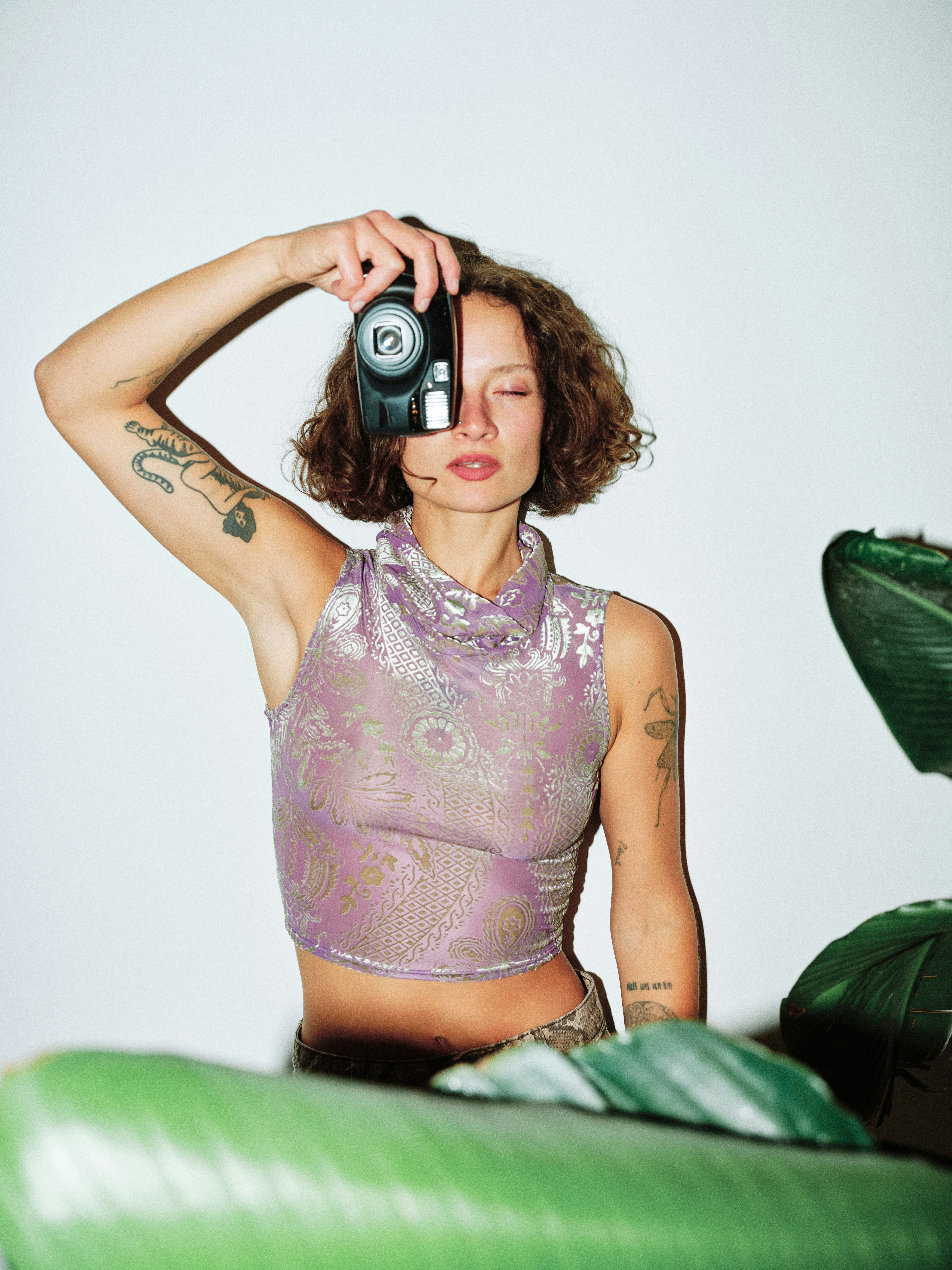 Editorial portrait of a tattooed model in a purple metallic crop top holding a film camera, surrounded by large tropical leaves, white studio background