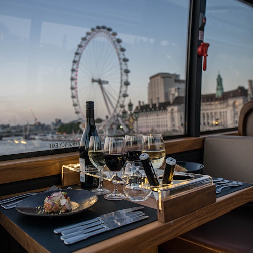 A table beside a window displays wine bottles, glasses, and a plated dish, overlooking a cityscape with a large Ferris wheel.