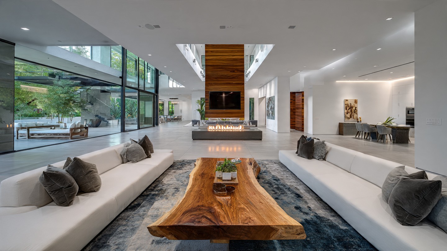Spacious modern living area with large glass windows, a central wood table, white sofas, and a sleek fireplace. Open, airy ambiance with natural light.