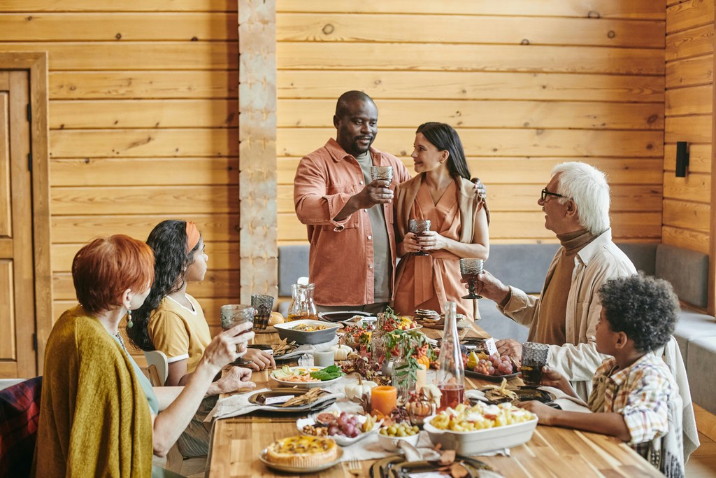 A diverse family enjoys a festive meal together in a cozy, wooden cabin, raising their glasses in a toast around a table filled with a variety of delicious foods.