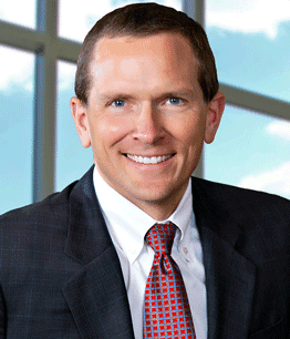 A smiling man in a suit is positioned in front of a bright window, with a blue sky visible outside.
