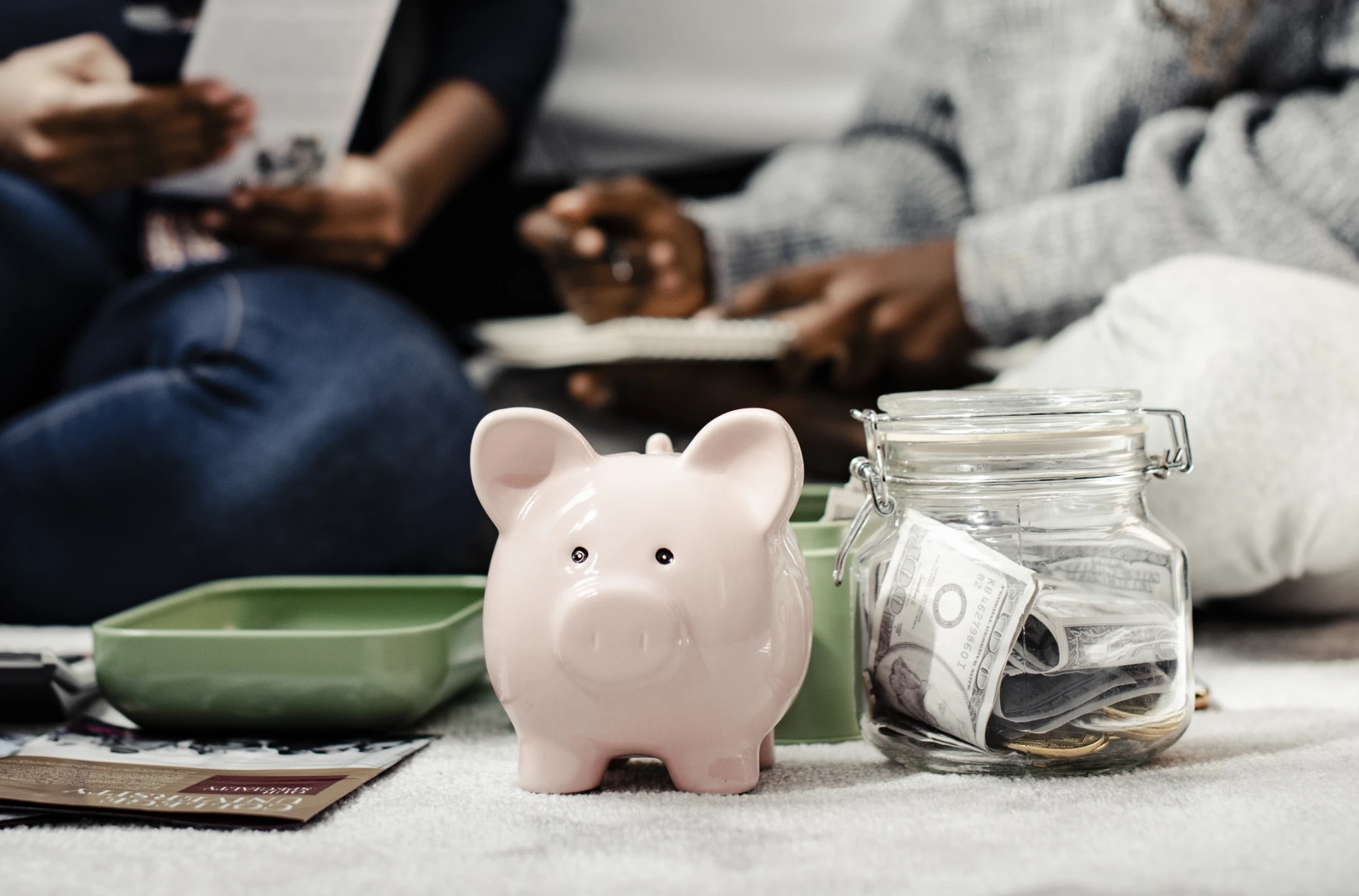 A pink ceramic piggy bank sits next to a glass jar filled with cash during a group financial planning meeting on a carpet.
