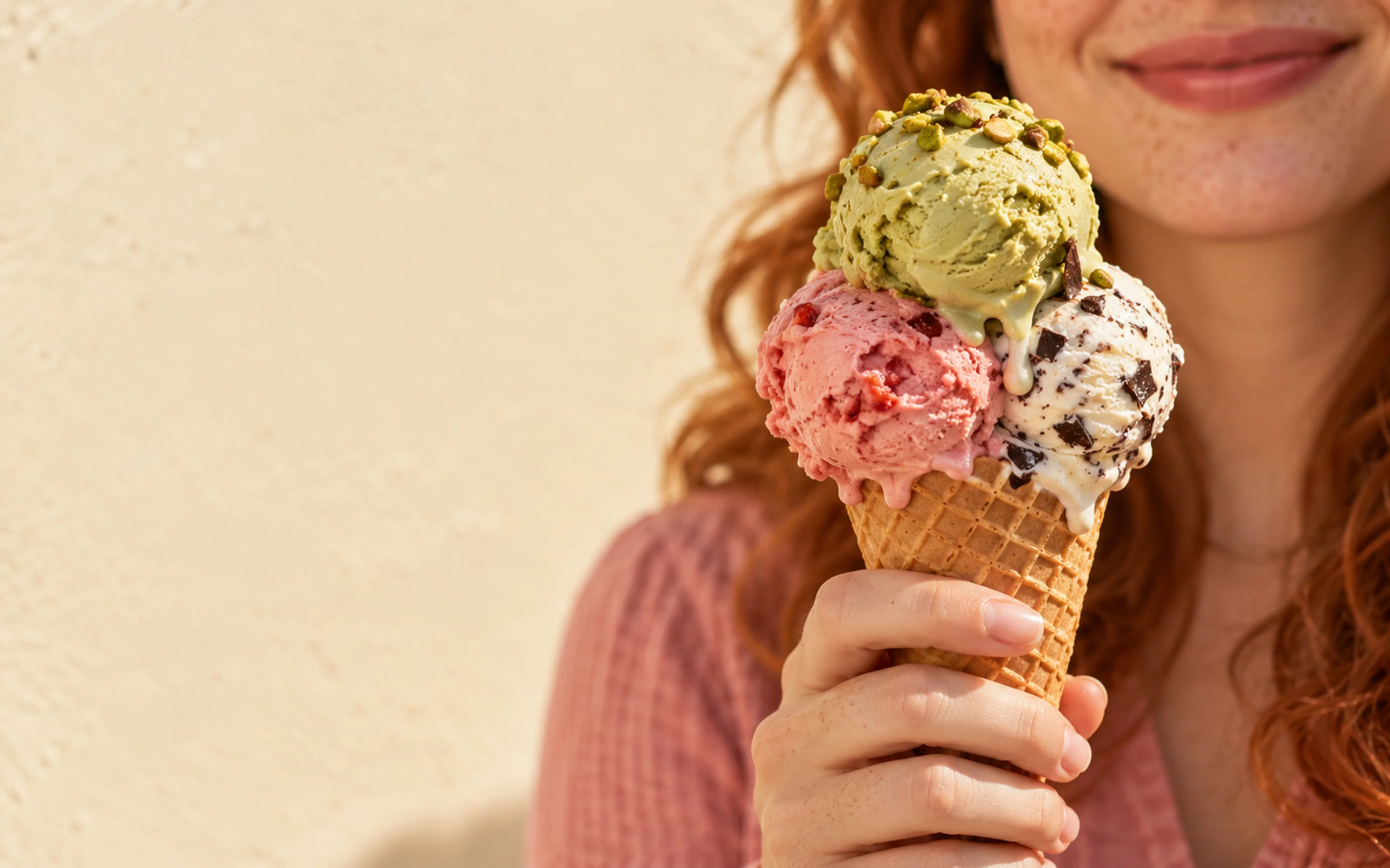 A three-scoop gelato cone of pistachio, strawberry and stracciatella held up in the sun