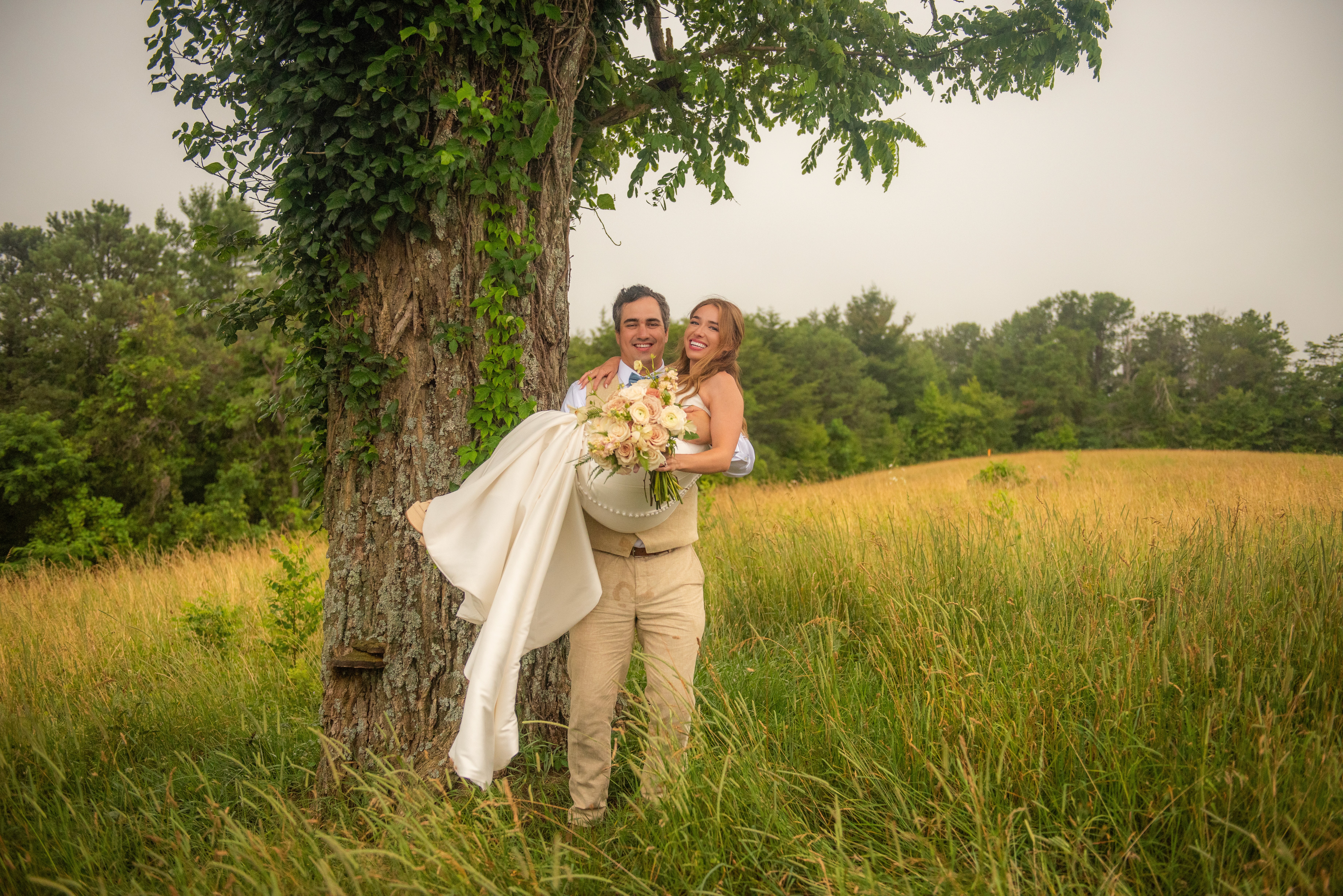 Happy groom holding his bride in a field during wedding day, smiling. She is holding white flowers. 