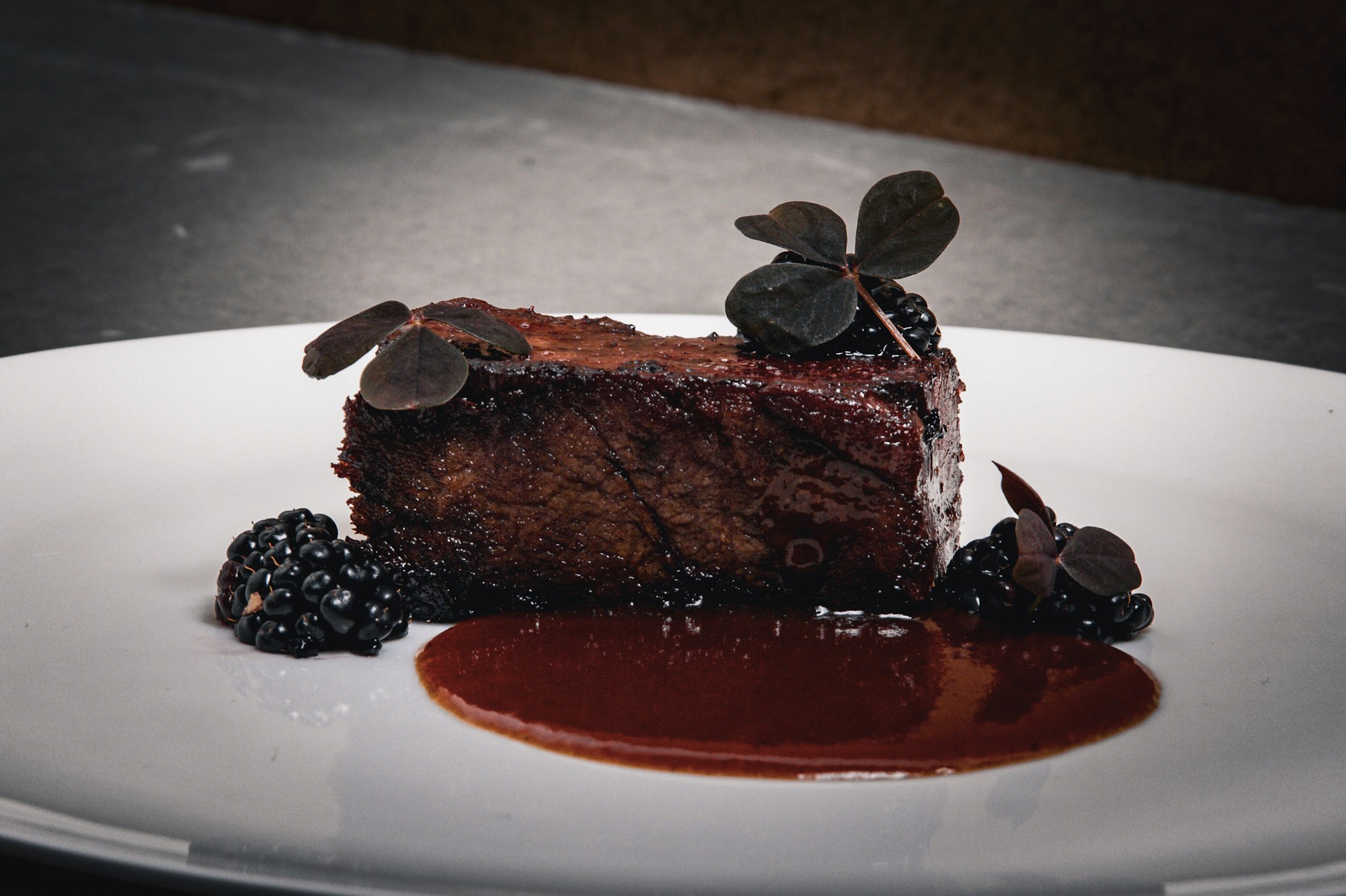 Close-up of glazed short rib with thick blackberry sauce, pickled blackberries, and sorrel on a white plate.