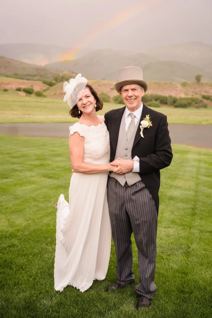 The bride and groom holding hands and smiling on a green lawn with a vibrant rainbow appearing in the sky behind them at a Chaparral Ranch Wedding.