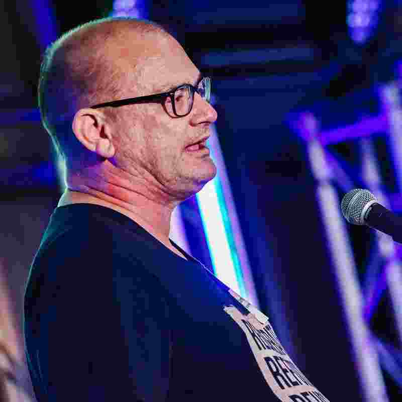 A man with short hair and glasses wearing a black t shirt stands in front of a microphone
