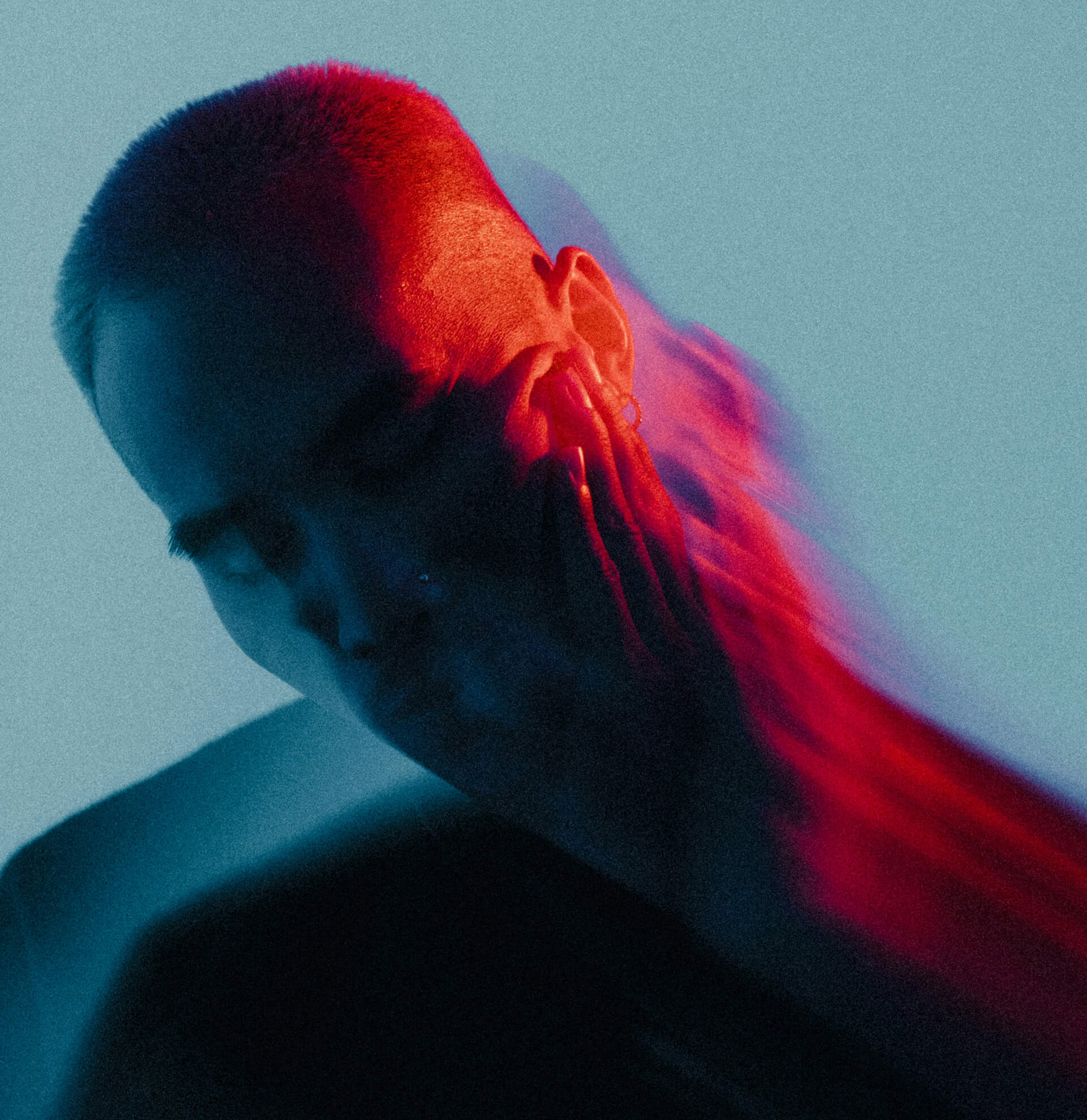 Portrait of a person with their eyes closed, hand on their face, and a blurred motion effect creating a trailing ghost-like silhouette. The subject is lit with vibrant red and blue lighting against a muted teal background, evoking a moody, introspective atmosphere.