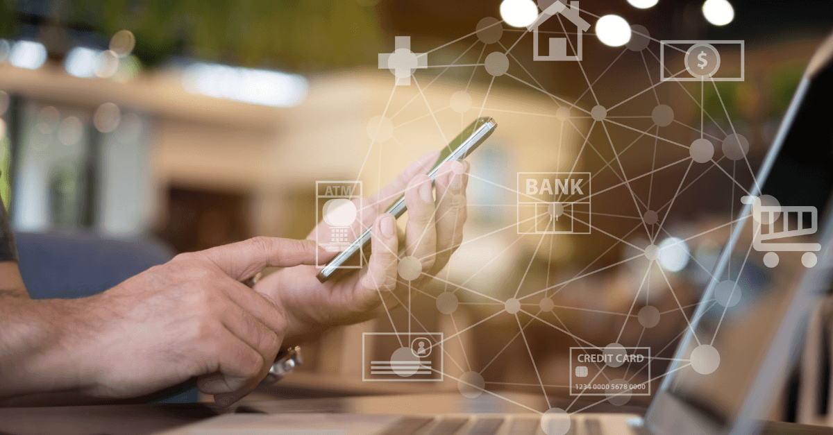 Closeup on a person's hands typing on a smartphone while a laptop sits on the table in front of them. A digital network overlay shows icons like home, bank, shopping cart, and credit card.