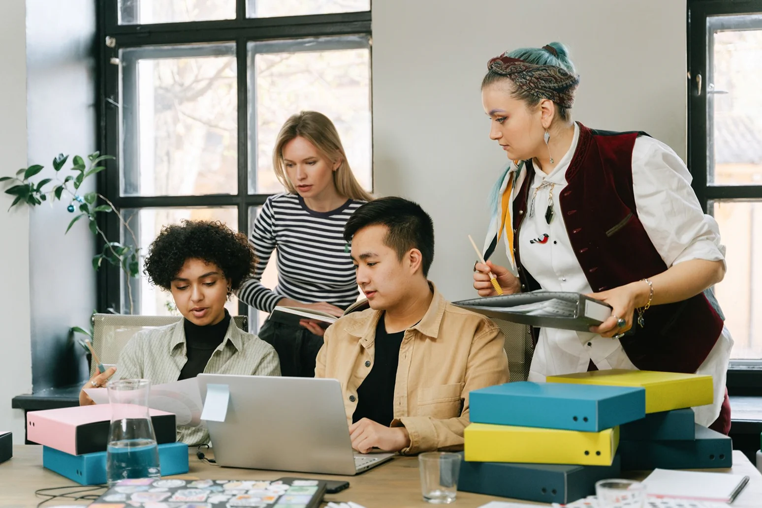A diverse group of four young creatives collaborating around a desk in a bright workshop, looking at a laptop and reviewing project documents.