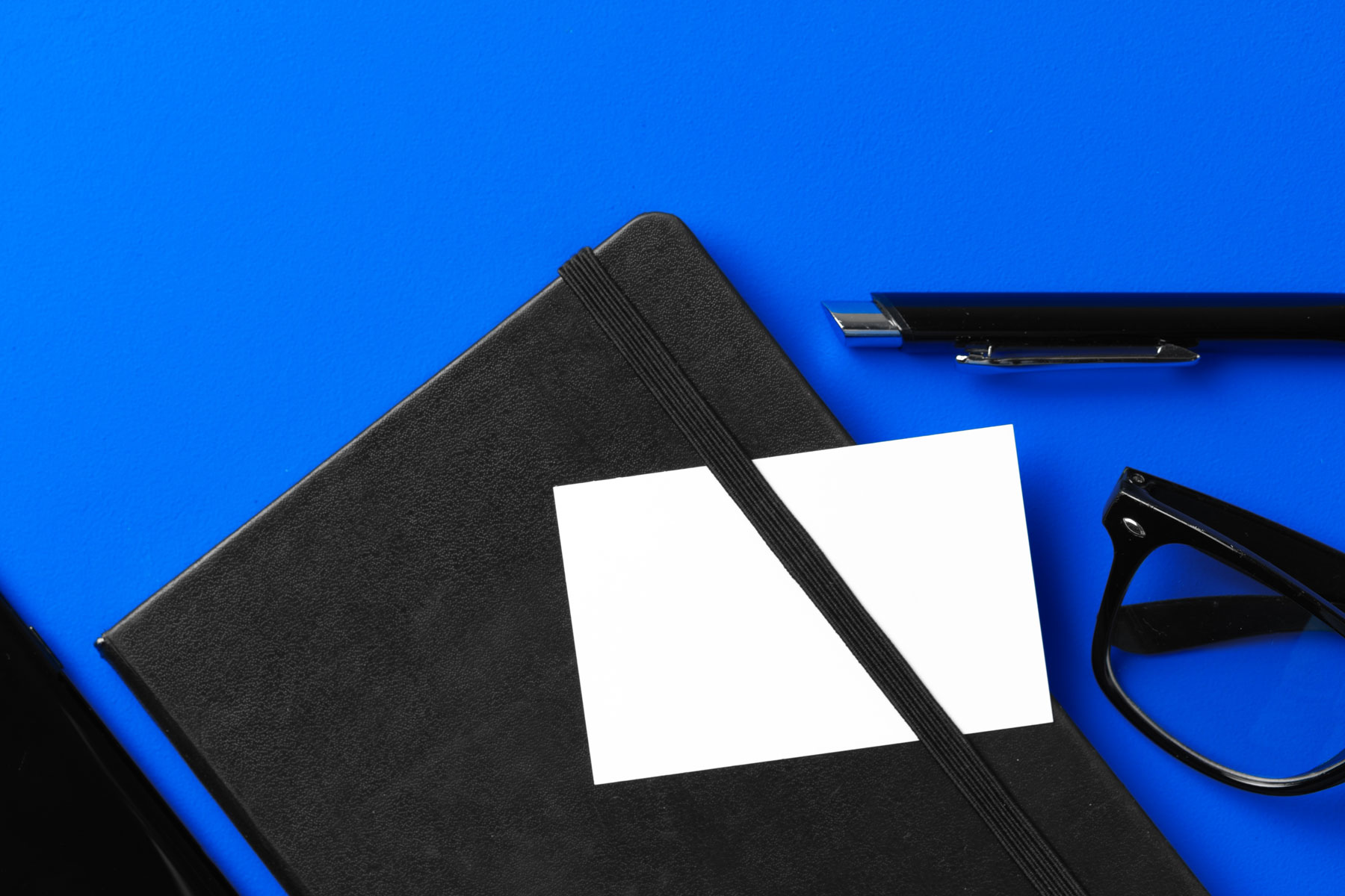 Biała pusta wizytówka umieszczona na czarnym eleganckim notesie obok okularów oraz pióra na jaskrawym niebieskim tle.