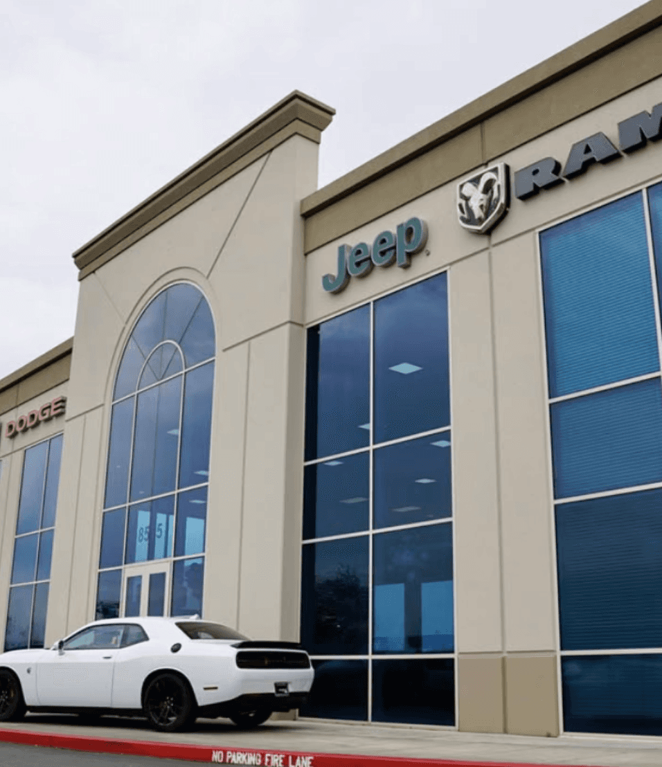 A rugged, green off-road vehicle is parked on gravel, flanked by other vehicles, featuring a prominent front grille and large tires under a clear blue sky.