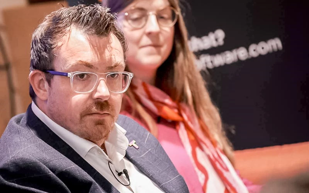 Two speakers are seated on stage during a live discussion, with event branding visible in the background. The lighting and close framing emphasise the speaker’s engagement.