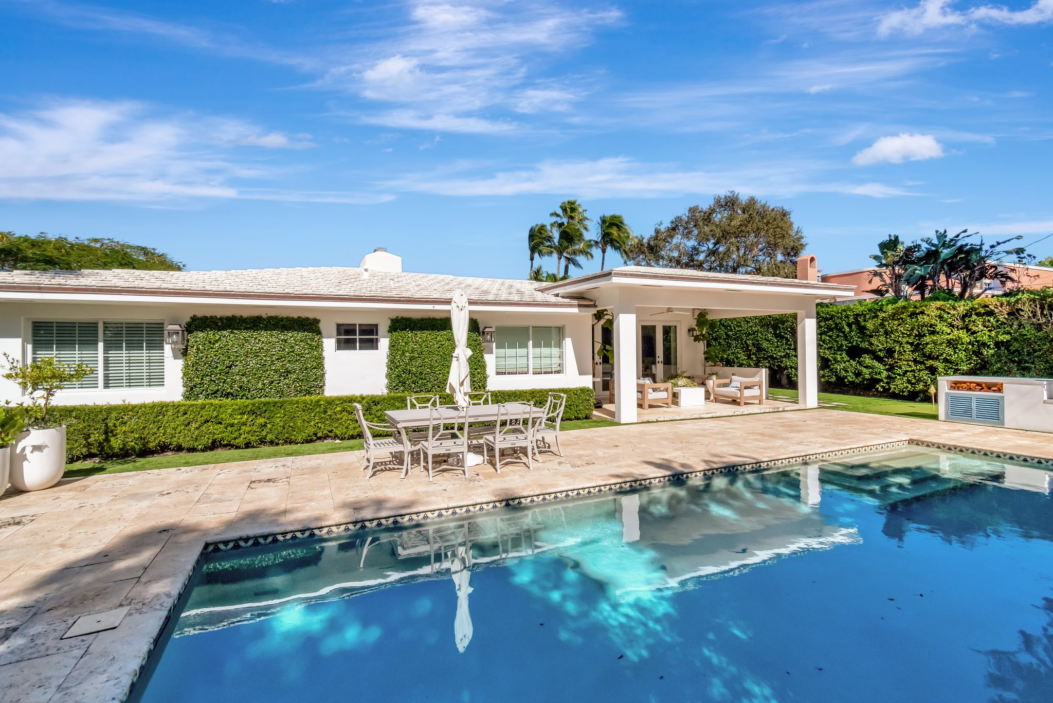 Single-story white Florida home with tropical landscaping, stone patio, outdoor dining set, and swimming pool reflecting palm trees under clear blue sky