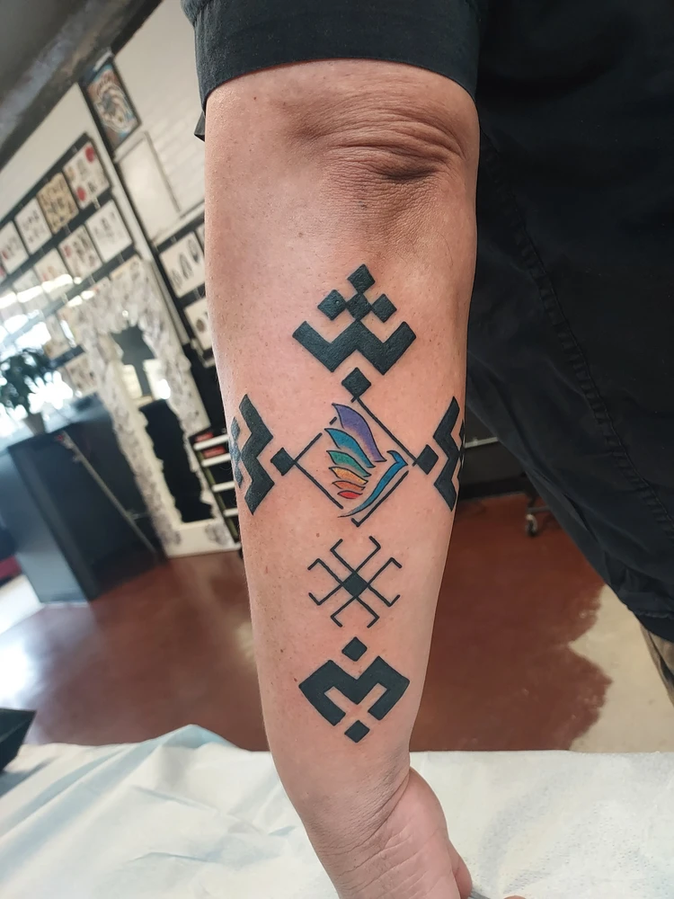 O antebraço de uma pessoa apresenta uma tatuagem geométrica em tinta preta com um tsuru de origami colorido no centro, cercada por padrões simétricos marcantes, contra o cenário de um estúdio de tatuagem com obras de arte emolduradas na parede.