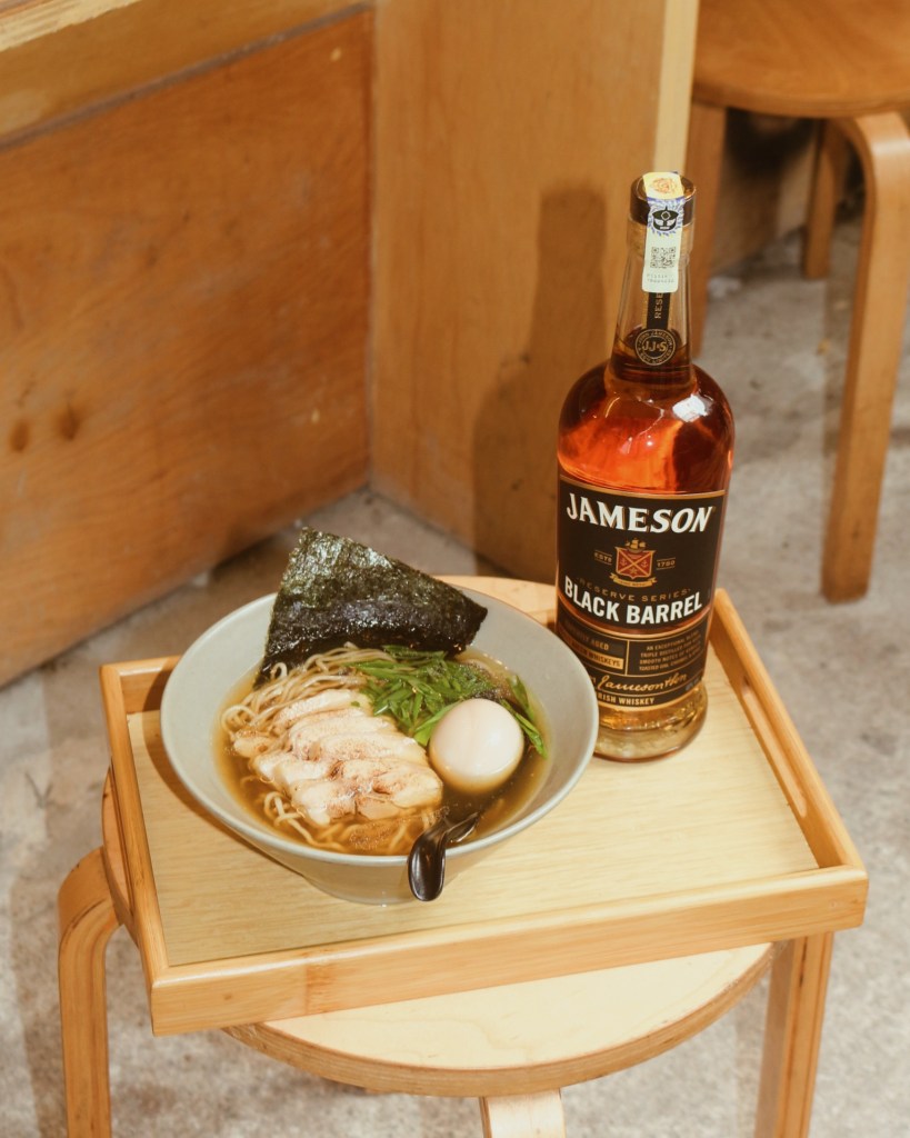 A bowl of chicken ramen with sliced chicken, a soft-boiled egg, a piece of seaweed, and a bottle of Jameson Black Barrel whiskey on a wooden tray.